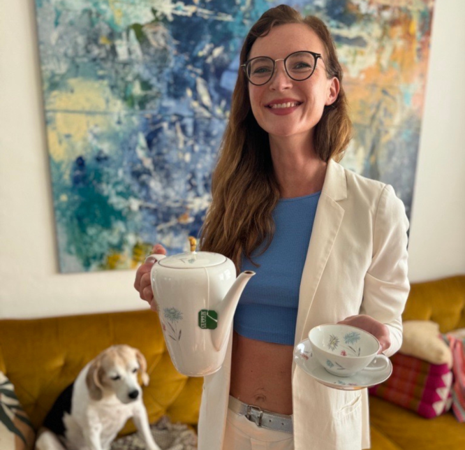 Eine Frau, die Ankerkopf Gründerin Sarah hält eine Kanne Tee in ihren Händen und schaut positiv in die Kamera. Dahinter sitzt ein Beagle auf einem Sofa. Dahinter befindet sich ein abstraktes Wandgemälde.